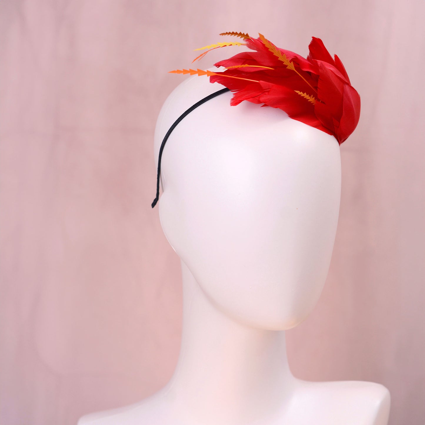 A bright red feather fascinator hat with orange-tipped feathers, displayed on a white mannequin head against a soft pink background.