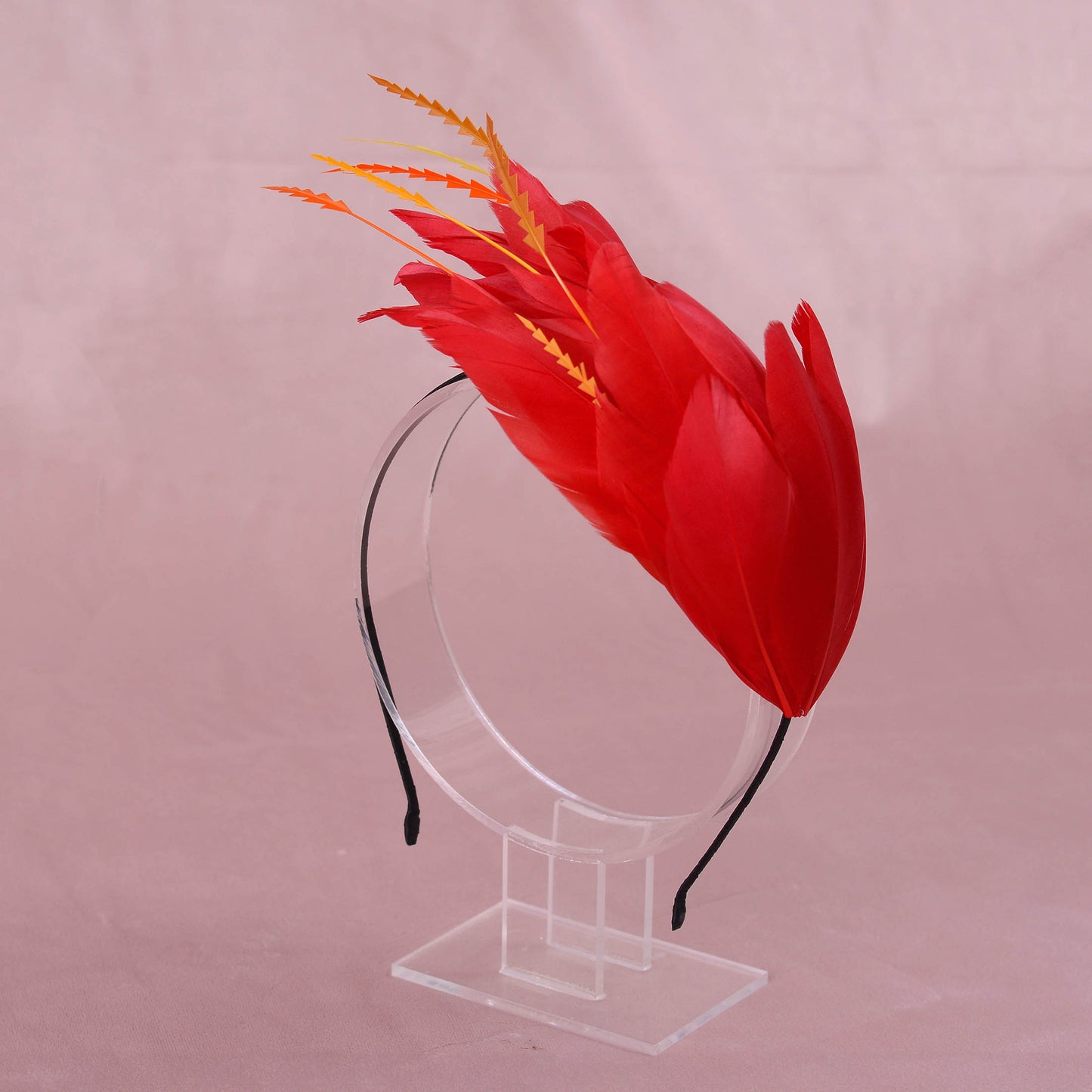 A red feather fascinator headband with orange and yellow accent feathers, displayed on a clear acrylic stand against a soft pink background.