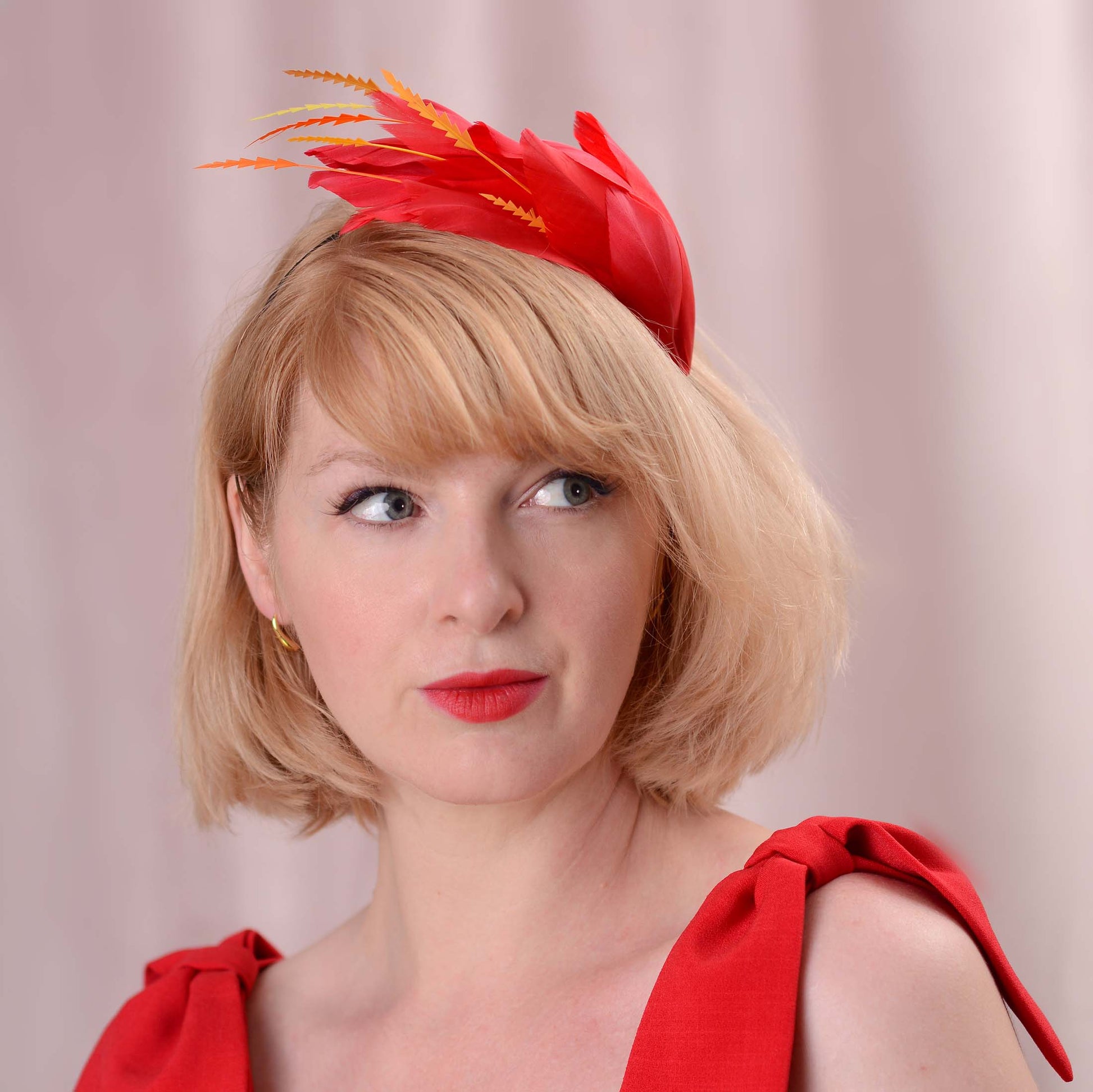 A woman with short blonde hair wearing a bright red feather fascinator with orange and yellow feather accents, paired with a matching red dress and lipstick against a soft pink background.