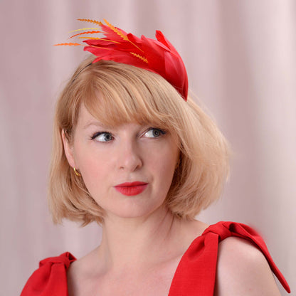 A woman with short blonde hair wearing a bright red feather fascinator with orange and yellow feather accents, paired with a matching red dress and lipstick against a soft pink background.