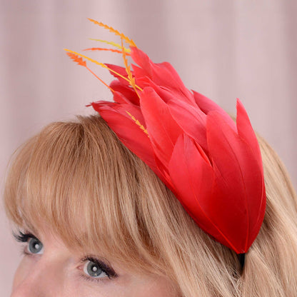 A close-up of a woman with blonde hair wearing a bright red feather fascinator headpiece with delicate orange and yellow feathers, against a soft pink background.