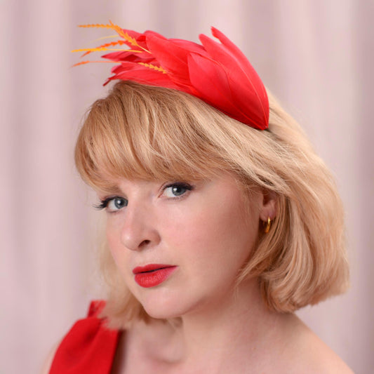 A close-up portrait of a woman with blonde hair wearing a bright red fascinator headpiece with orange feather accents, against a soft pink backdrop.