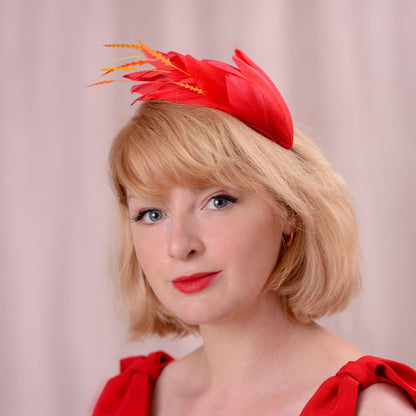 A woman with short blonde hair wearing a bright red fascinator hat with orange feather accents, posed against a soft pink studio backdrop.