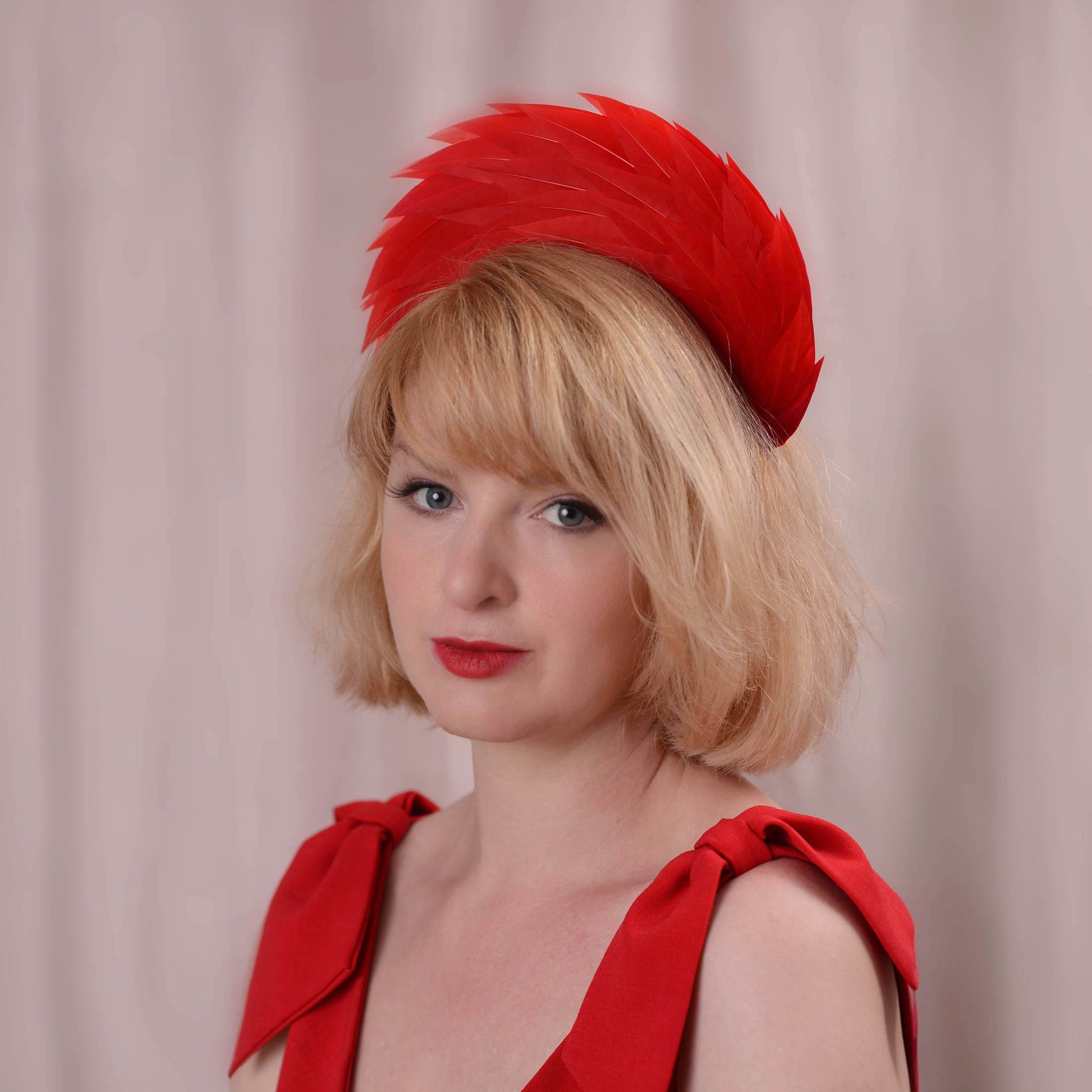 A blonde woman wearing a bright red feathered fascinator headband and a matching red dress photographed against a soft pink curtain background.