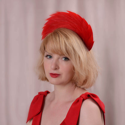 A blonde woman wearing a bright red feathered fascinator headband and a matching red dress photographed against a soft pink curtain background.
