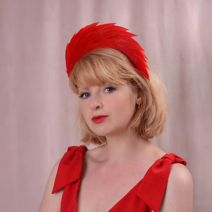 A woman with a blonde bob wearing a bright red feathered fascinator headband and a matching red dress photographed against a soft pink curtain background.