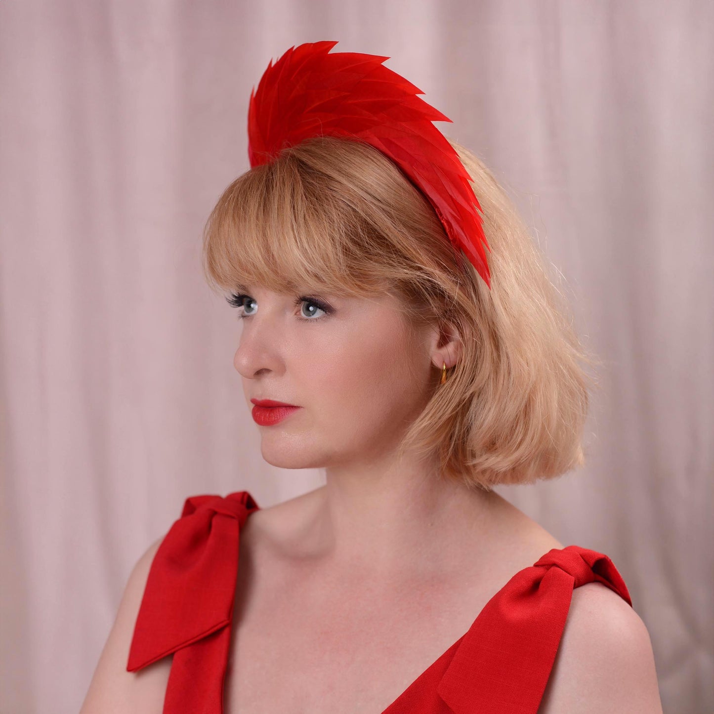 A woman with blonde hair wearing a bright red feather fascinator headband and a matching red top, photographed against a soft pink curtain background.