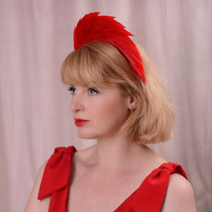 A woman with blonde hair wearing a bright red feather fascinator headband and a matching red top, photographed against a soft pink curtain background.
