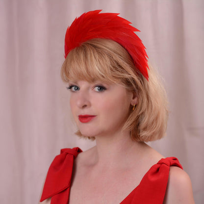A woman with short blonde hair wearing a bright red feather fascinator headpiece and a matching red bow-front dress, photographed against a soft pink curtain backdrop.