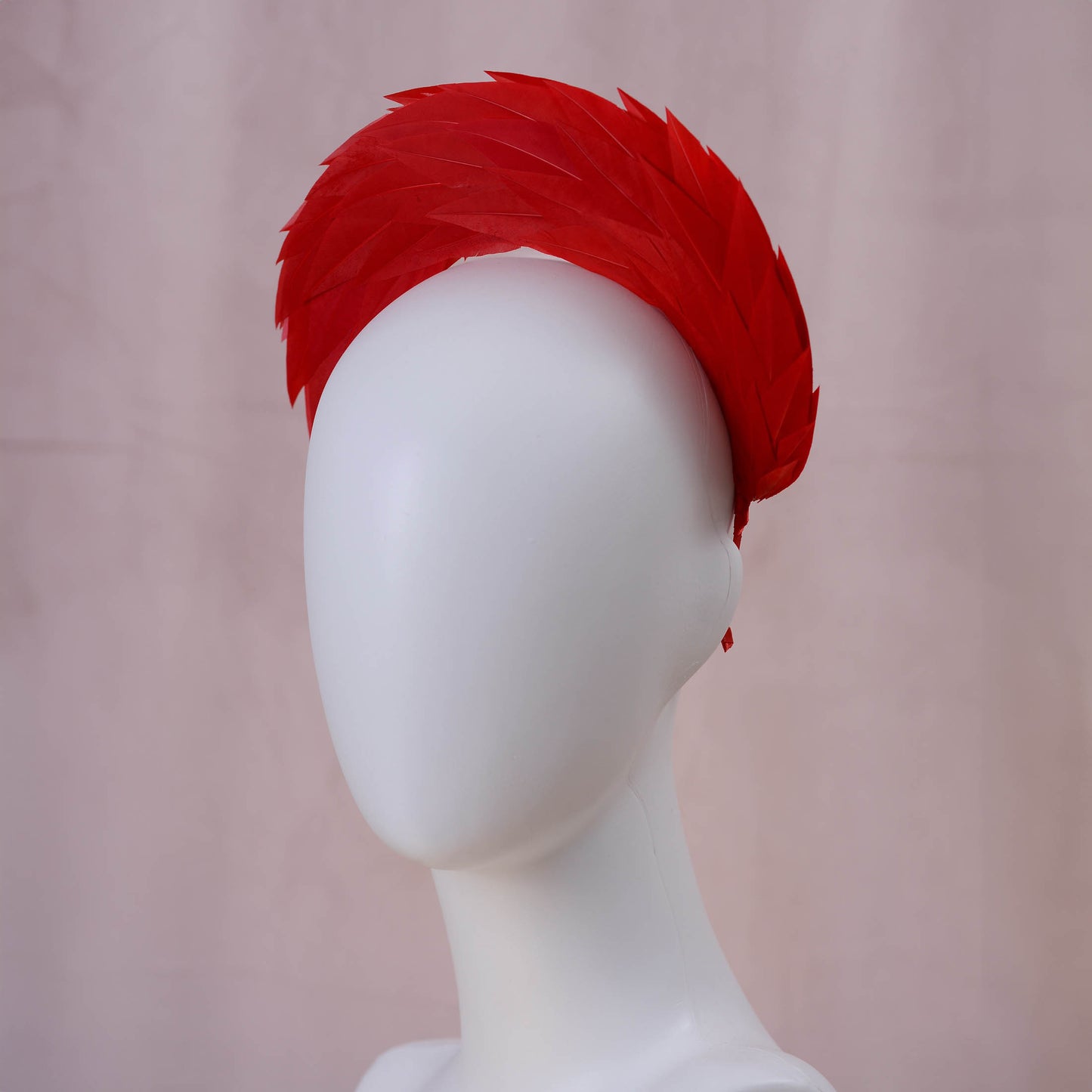 A bright red feather headband displayed on a white mannequin head against a soft pink backdrop.