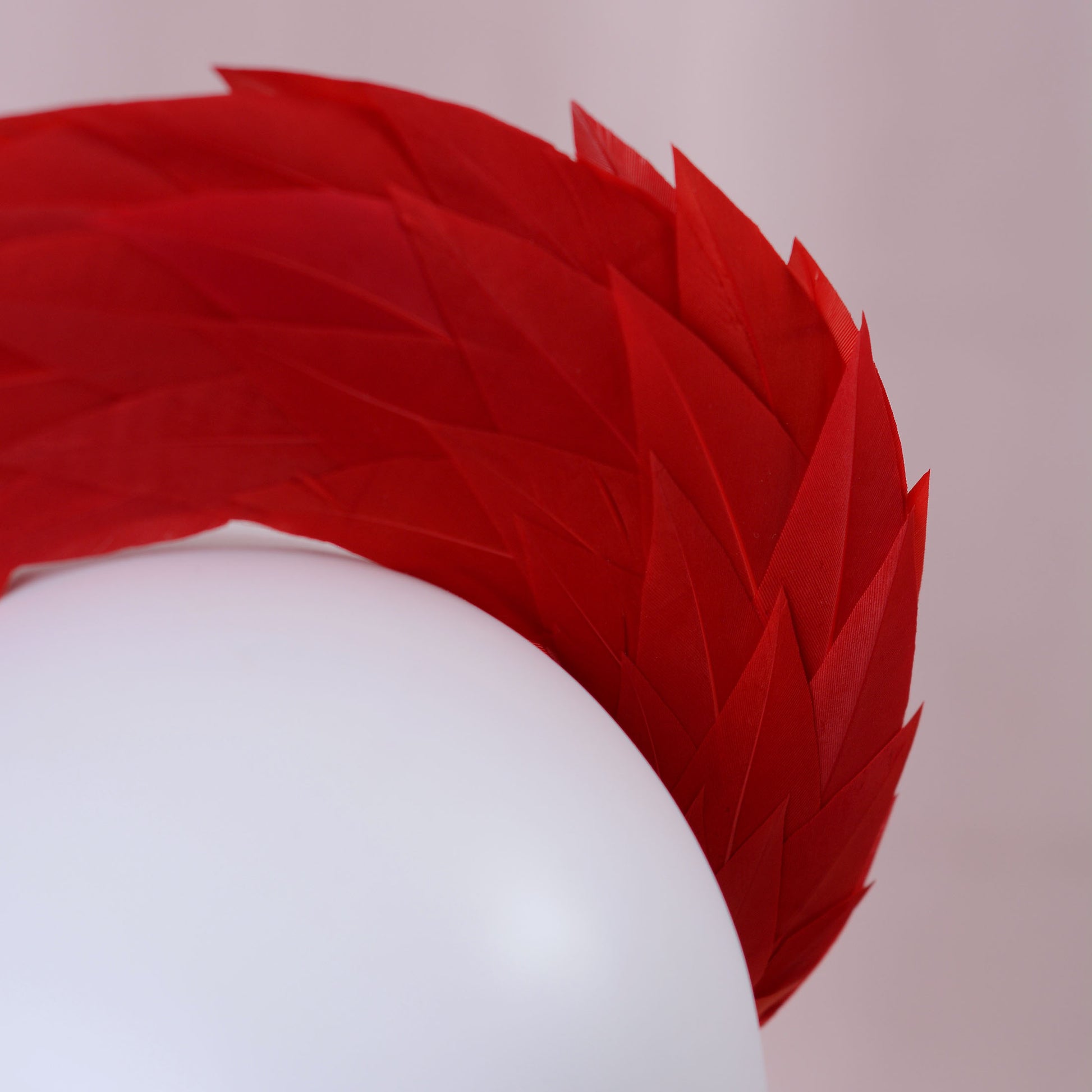 A close-up of a bright red layered feather headband accessory displayed on a white mannequin head against a soft light background.