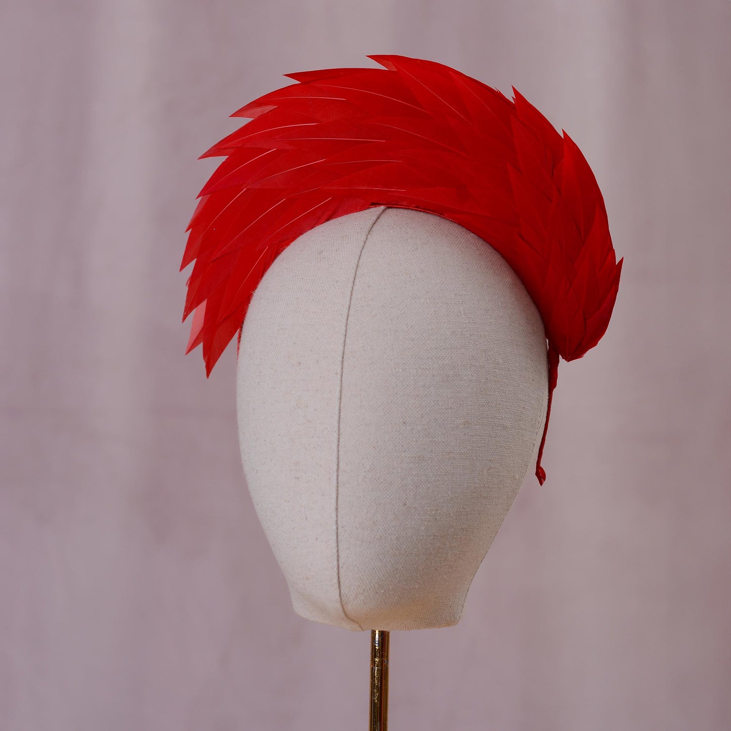 A bright red feather headband styled in a high halo displayed on a white fabric mannequin head against a soft, light pink background front on view.