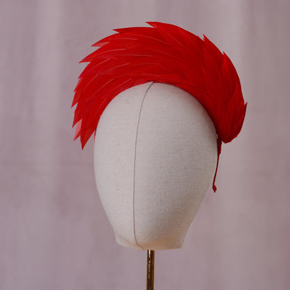A bright red feather headband styled in a high halo displayed on a white fabric mannequin head against a soft, light pink background front on view.
