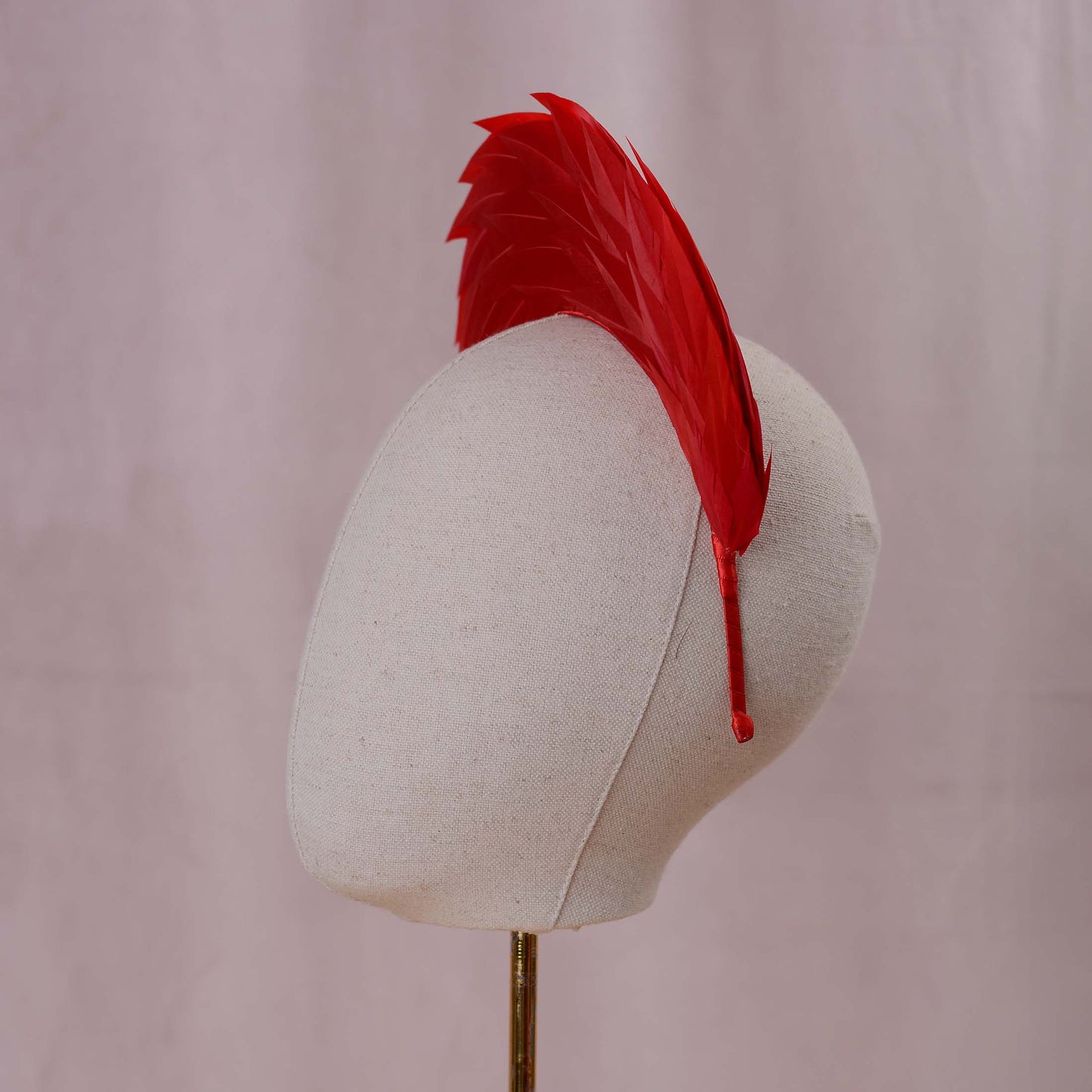 A bright red feather headband styled in a high halo displayed on a white fabric mannequin head against a soft, light pink background side on view.