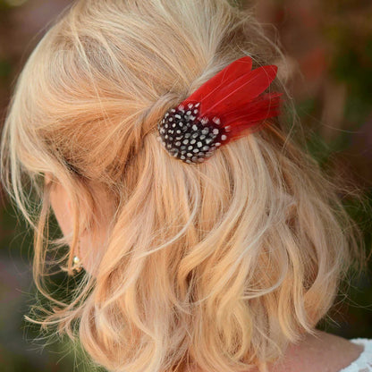A close up of  blonde woman shown from the back wearing a short, wavy half up hair style with a red feather hair accessory. The accessory features a black and white polka dot guinea fol feathers with bright red feathers. 