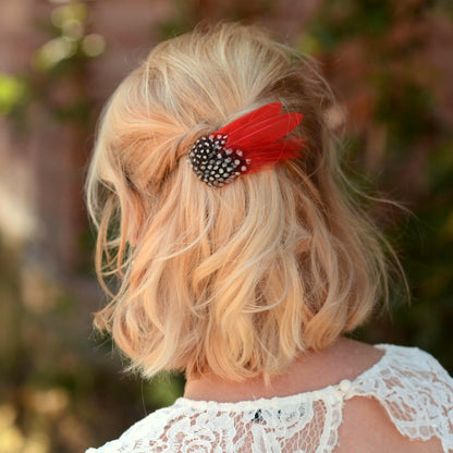 A blonde woman shown from the back wearing a short, wavy half up hair style with a red feather hair accessory. The accessory features a black and white polka dot guinea fol feathers with bright red feathers. 