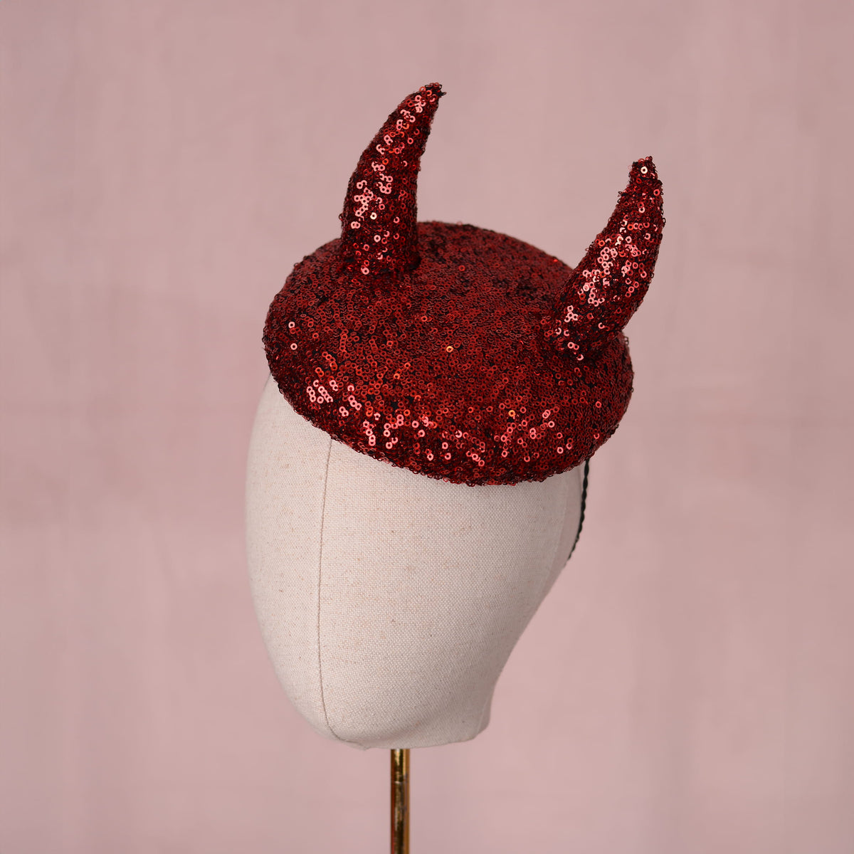 A red sequin beret hat with two 3D devil horns displayed on a cream fabric mannequin head against a soft pink background.