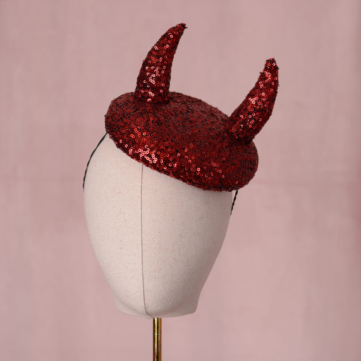 A red sequin beret hat with two 3D devil horns displayed on a cream fabric mannequin head against a soft pink background.