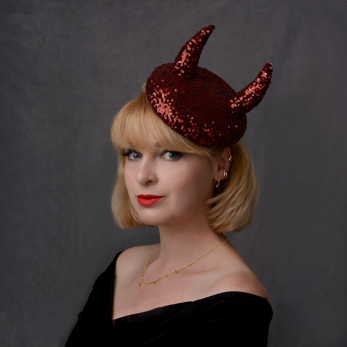 A woman with short blonde hair models a dark red sequin devil horned beret hat against a dark gray studio background.