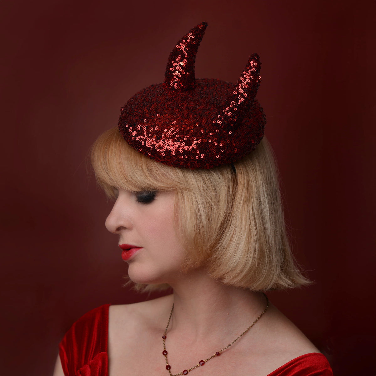 A woman with a short blonde bob wears a red sequin hat with two 3d devil horns,  in profile posed against a dark red background.