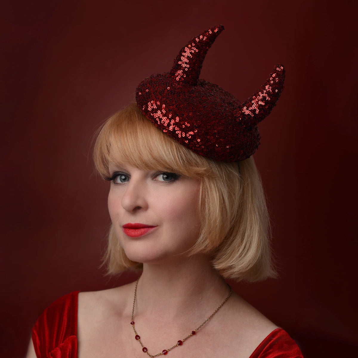 A woman with a short blonde bob wears a red sequin devil horn beret headpiece against a dark red studio background.