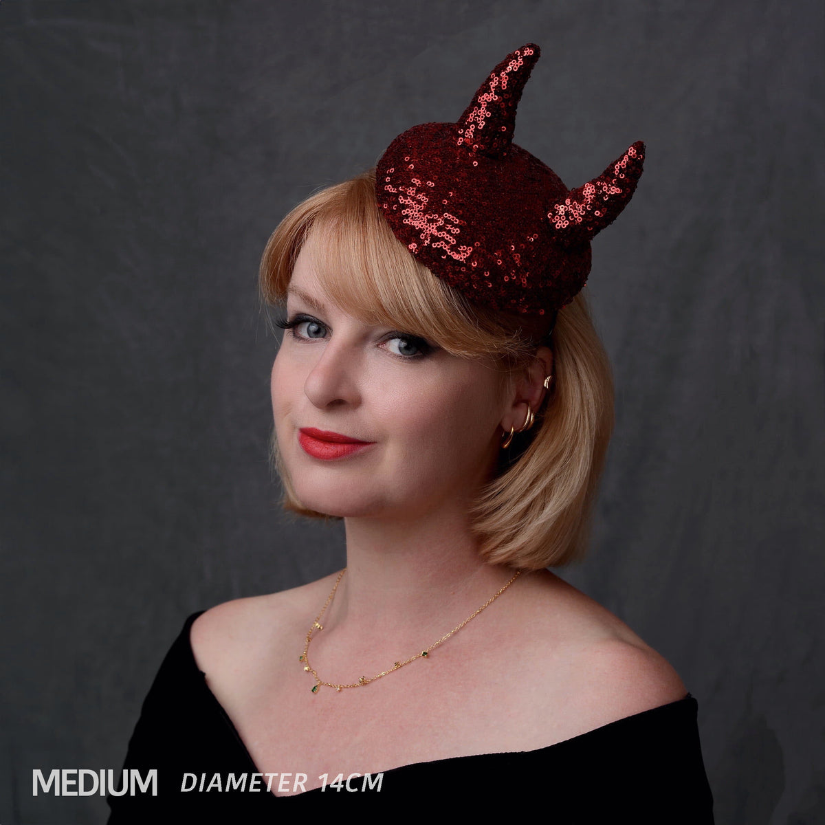 A woman with blonde hair wearing a small red sequin devil horn hat against a dark gray background. Text in the bottom left reads 'MEDIUM DIAMETER 14CM'.