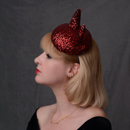 A woman in profile wearing a sparkling red sequin devil horn button fascinator hat against a dark gray background.