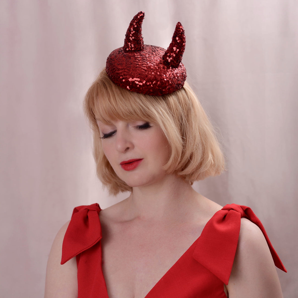 A woman with blonde hair wearing a red sequin side perching button hat with two 3D devil horns, paired with a red dress, against a soft pinkish-beige background.