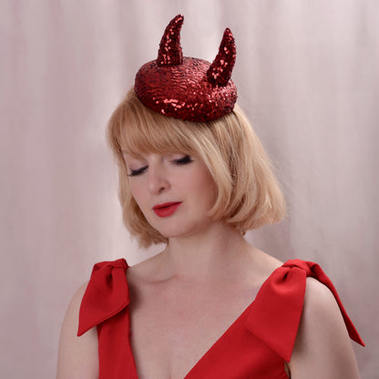 A woman with blonde hair wearing a red sequin side perching button hat with two 3D devil horns, paired with a red dress, against a soft pinkish-beige background.