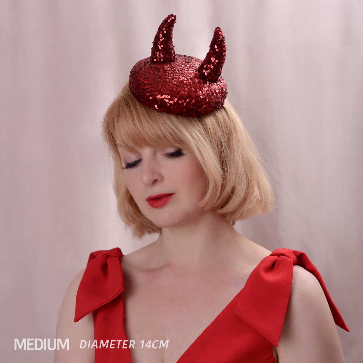 A woman with short blonde hair models a red sequined devil hat with two 3d horns, wearing a red dress with large shoulder bows. The text 'MEDIUM DIAMETER 14CM' is visible in the bottom-left corner.