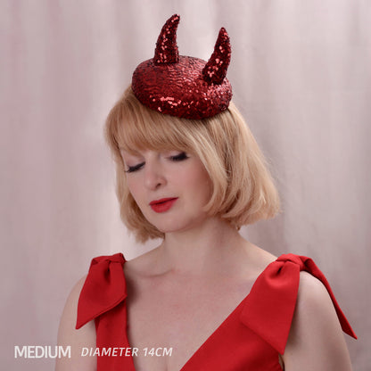 A woman with short blonde hair models a red sequined devil hat with two 3d horns, wearing a red dress with large shoulder bows. The text 'MEDIUM DIAMETER 14CM' is visible in the bottom-left corner.