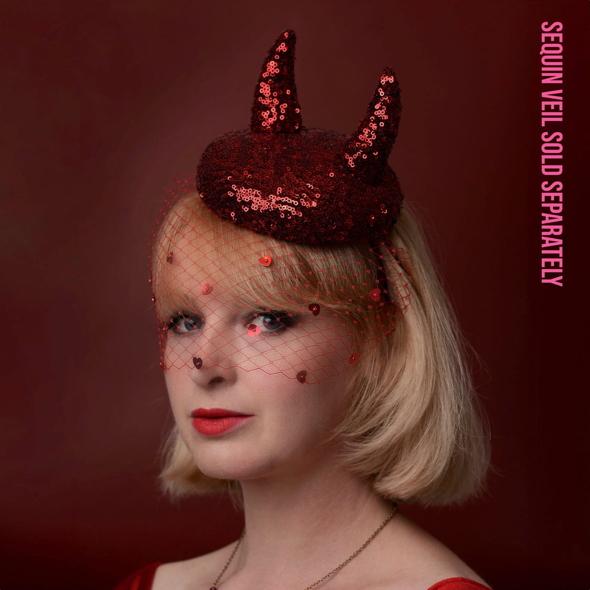 A woman with short blonde hair wears a dark red sequin horned headpiece with a matching red netted veil with sequin hearts. The background is a solid dark red, and pink vertical text on the right reads 'SEQUIN VEIL SOLD SEPARATELY'.