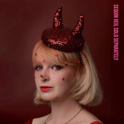 A woman with short blonde hair wears a dark red sequin horned headpiece with a matching red netted veil with sequin hearts. The background is a solid dark red, and pink vertical text on the right reads 'SEQUIN VEIL SOLD SEPARATELY'.