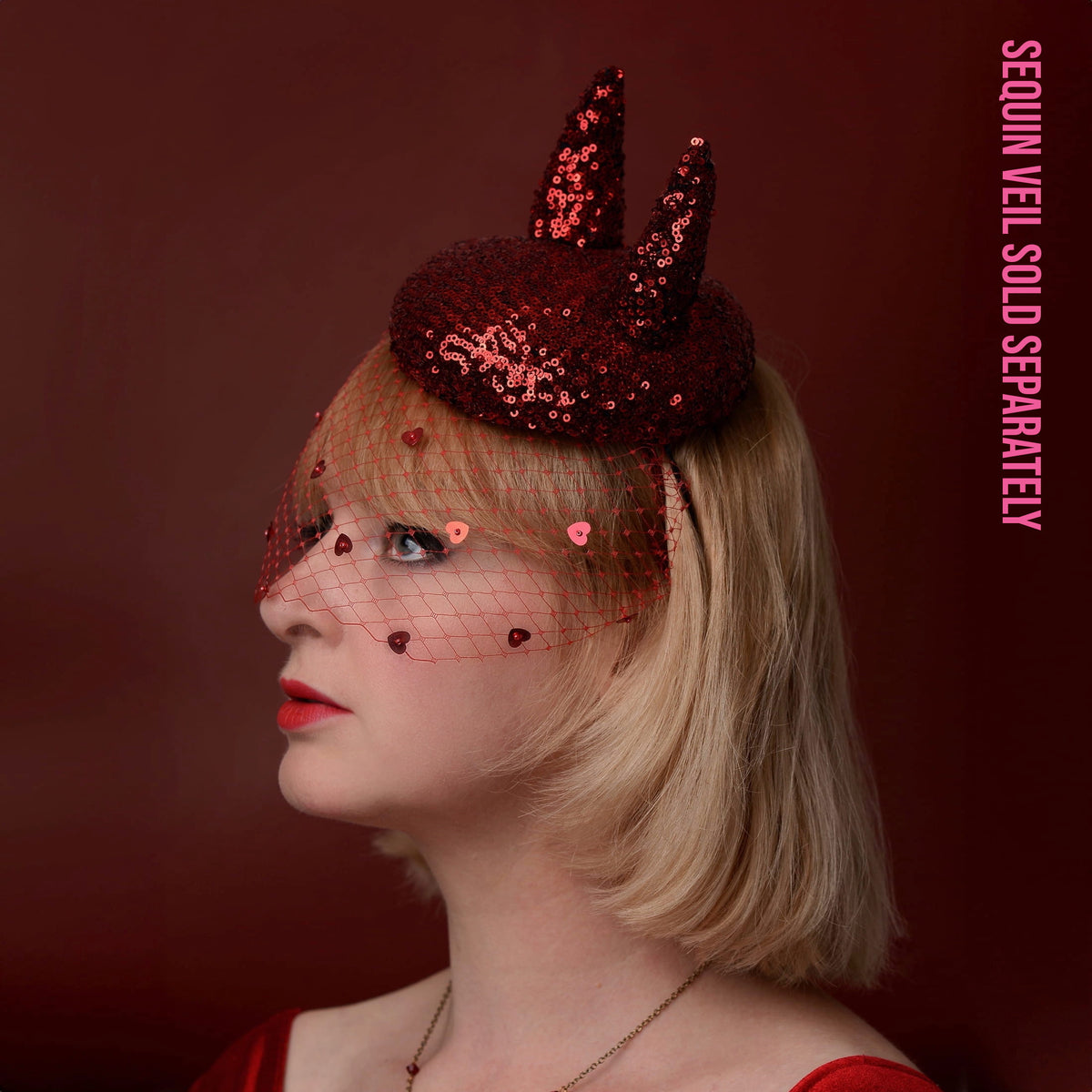 A woman in profile wearing a dark red sequin devil horned fascinator with a matching red sequin heart birdcage net veil. The background is a solid dark red, and pink vertical text on the right reads 'SEQUIN VEIL SOLD SEPARATELY'.