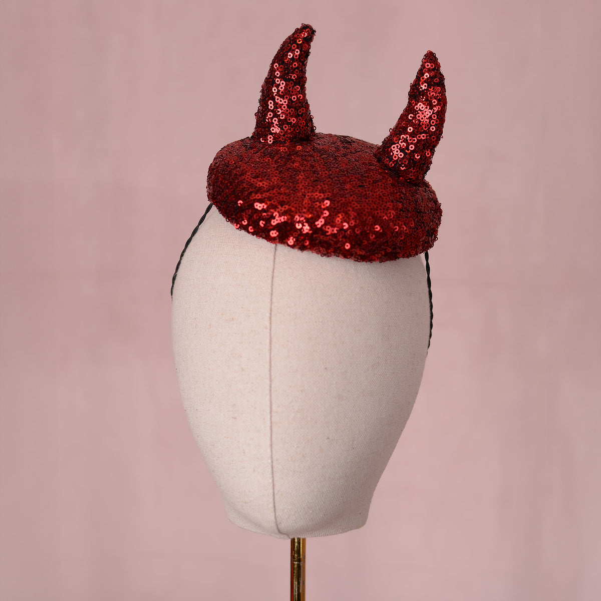 A red sequin horned  button shaped fascinator headpiece displayed on a cream fabric mannequin head against a soft pink background.
