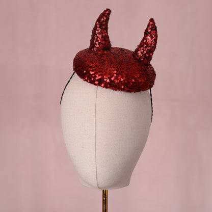 A red sequin horned  button shaped fascinator headpiece displayed on a cream fabric mannequin head against a soft pink background.