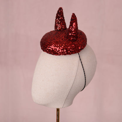 A red sequin button fascinator with 3D devil horns displayed on a cream fabric mannequin head against a soft pink background. A glamorous Halloween hat.