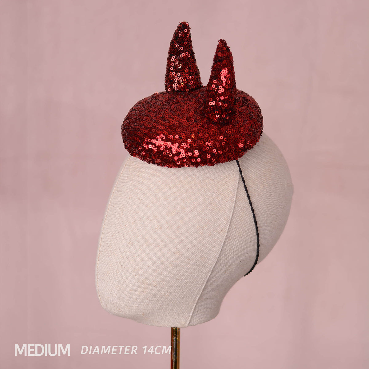 A red sequin devil horn mini hat displayed on a cream fabric mannequin head against a soft pink background, with text in the lower left reading 'MEDIUM DIAMETER 14CM'.