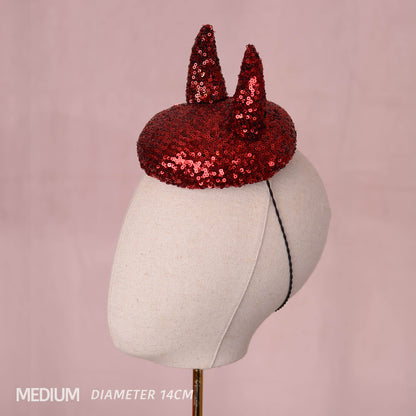 A red sequin devil horn mini hat displayed on a cream fabric mannequin head against a soft pink background, with text in the lower left reading 'MEDIUM DIAMETER 14CM'.
