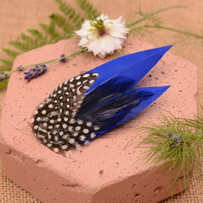 Blue feather hair clip with black and white polka dot design on a pink stone surface with flowers and plants.