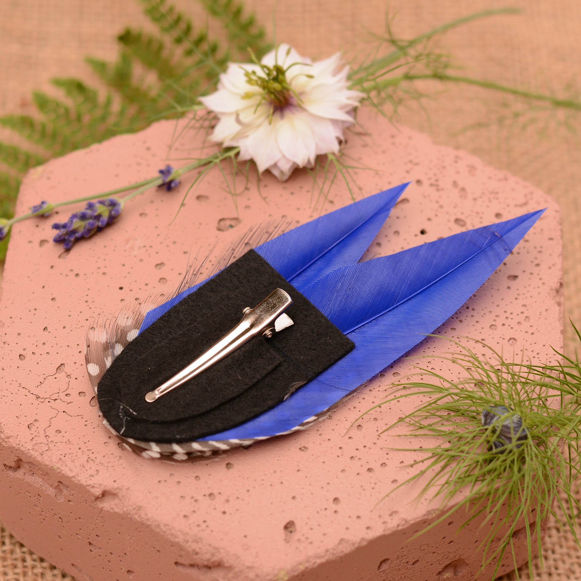 Blue feather hair clip with black and white polka dot design on a pink stone surface with flowers and plants.