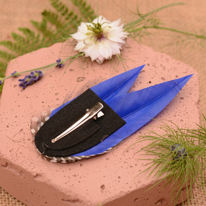 Blue feather hair clip with black and white polka dot design on a pink stone surface with flowers and plants.