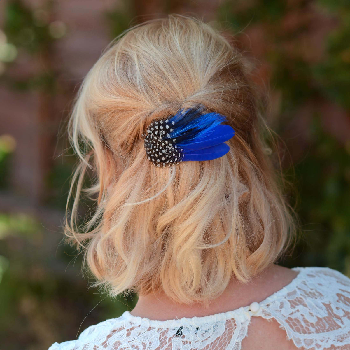 A woman with wavy blonde hair styled in a half up do  secured with a decorative hair clip featuring vivid royal blue feathers and black-and-white polka dot guinea fowl feathers