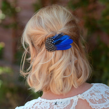 A woman with wavy blonde hair styled in a half up do  secured with a decorative hair clip featuring vivid royal blue feathers and black-and-white polka dot guinea fowl feathers
