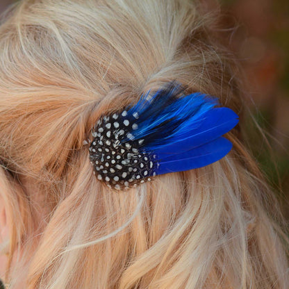 A close-up of blonde hair secured with a decorative hair clip featuring vivid royal blue feathers and black-and-white polka dot guinea fowl feathers
