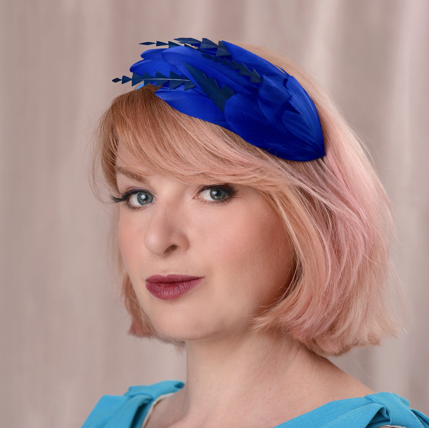 A woman with soft pink hair wearing a bright royal blue feather fascinator headpiece with navy blue details, set against a soft beige curtain background.