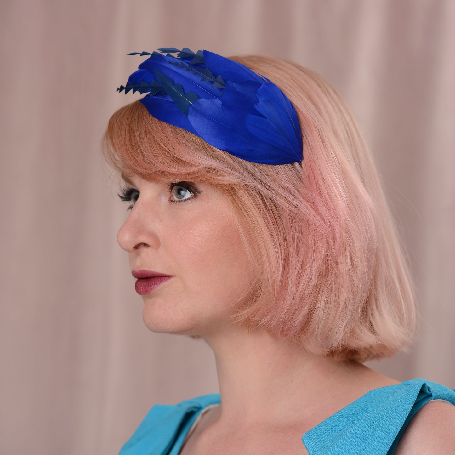 A woman with soft pink hair wearing a bright royal blue feather fascinator headpiece with navy blue details, set against a soft beige curtain background.
