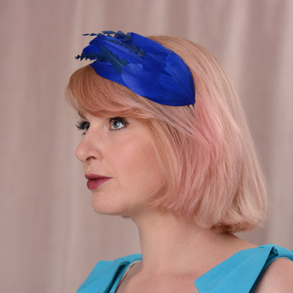 A woman with soft pink hair wearing a bright royal blue feather fascinator headpiece with navy blue details, set against a soft beige curtain background.