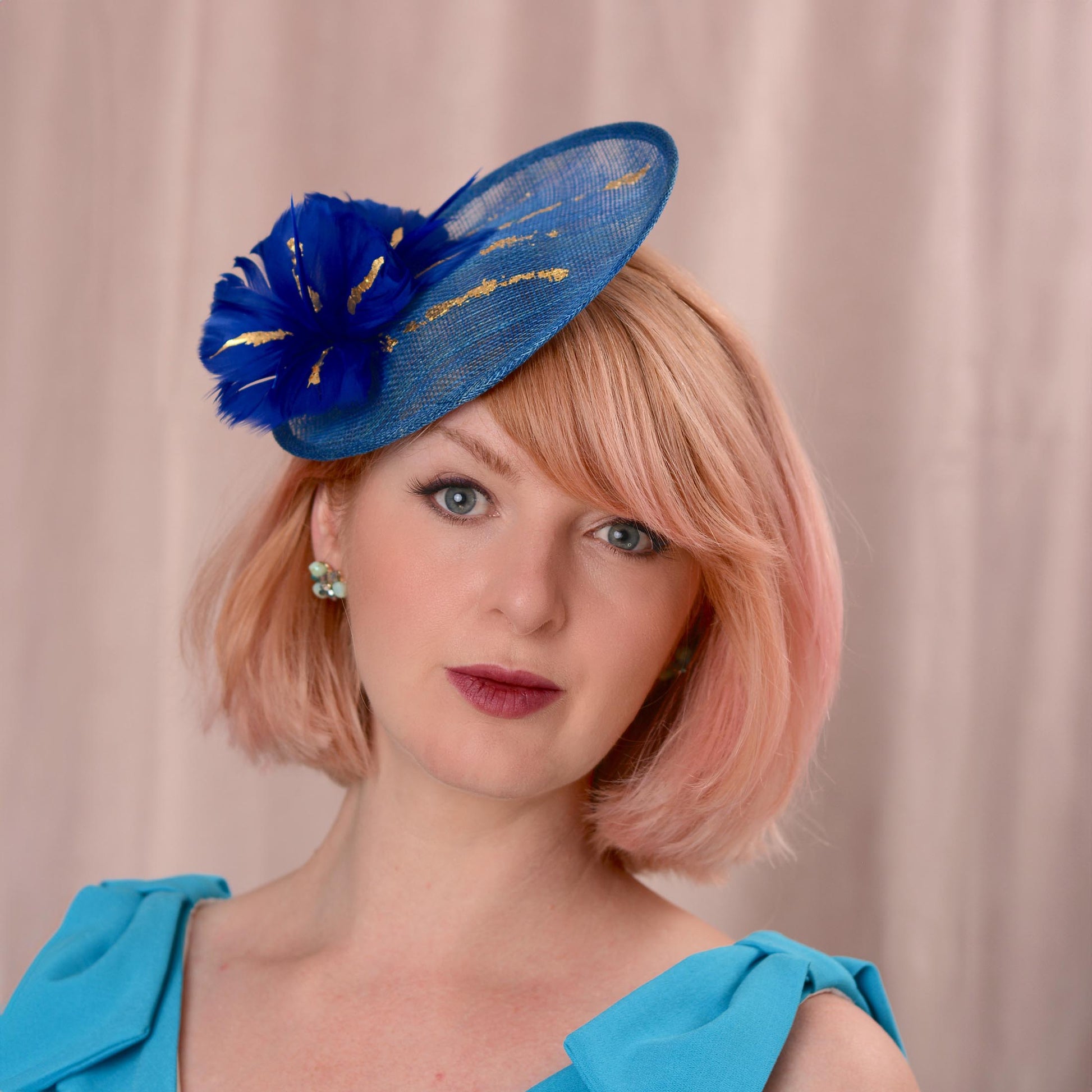 A woman with short pink hair wearing a royal blue fascinator hat with gold accents and a matching blue top, photographed against a soft beige curtain background.