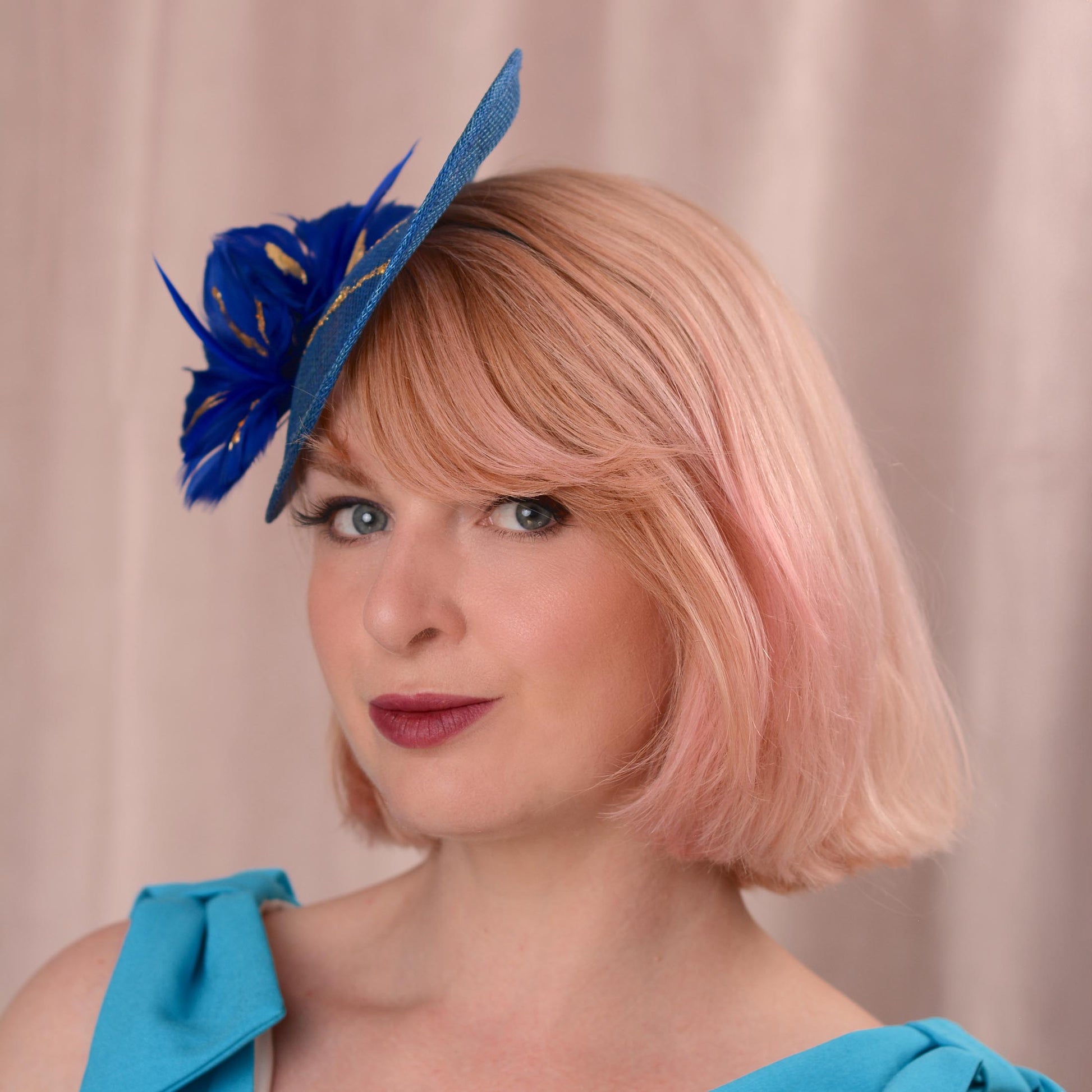 A woman with pink hair wearing a blue fascinator hat and a bright blue top, photographed against a soft beige curtain background.